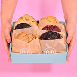 Hands holding 6 x New York Cookies, The BIG Birthday Box with assorted flavors in a blue gift box against pink background.